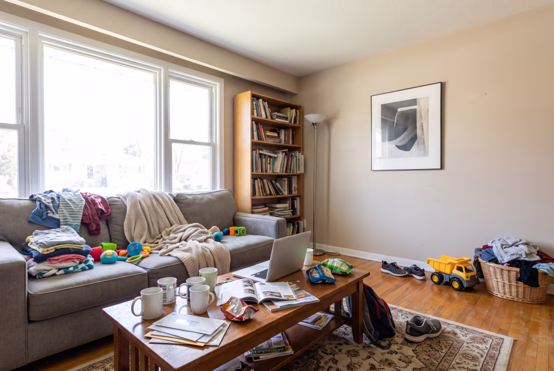 A cluttered living room with beige walls: laundry piled on a gray sofa beside baby toys, mugs, a laptop, snacks, magazines and a stack of mail on the coffee table, sneakers, a backpack and a toy dump truck on the floor, and an overflowing laundry basket against the wall.